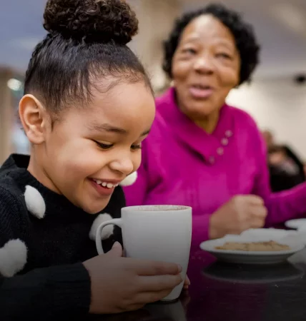 Young girl looking excitedly inside her mug at a hot chocolate, their guardian smiles next to her